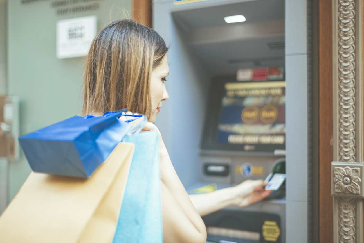 A woman holding shopping bags inserts a card into an ATM, preparing to withdraw cash. The scene is slightly blurred, focusing on her actions at the machine.