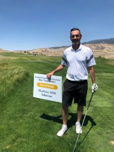A man in a white polo shirt and black shorts stands on a golf course, holding a golf club in one hand and a sign in the other. The sign promotes a company called MaxEffort ATM Solutions. Blue sky and hills are in the background.