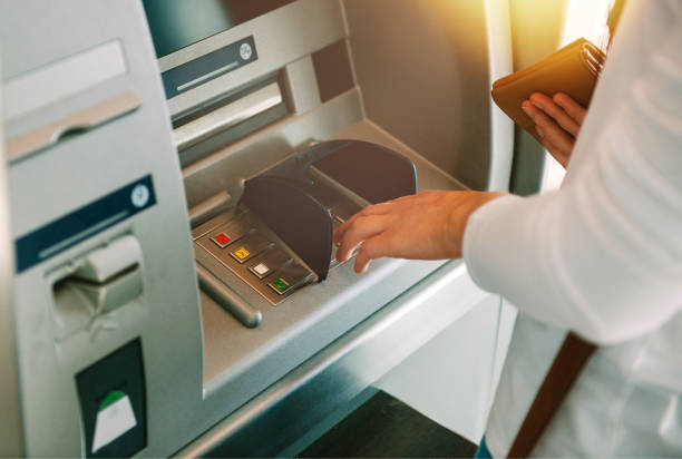 A business owner uses a cash machine, entering their PIN on the keypad with one hand while holding a wallet in the other, as sunlight streams in from the top right corner.