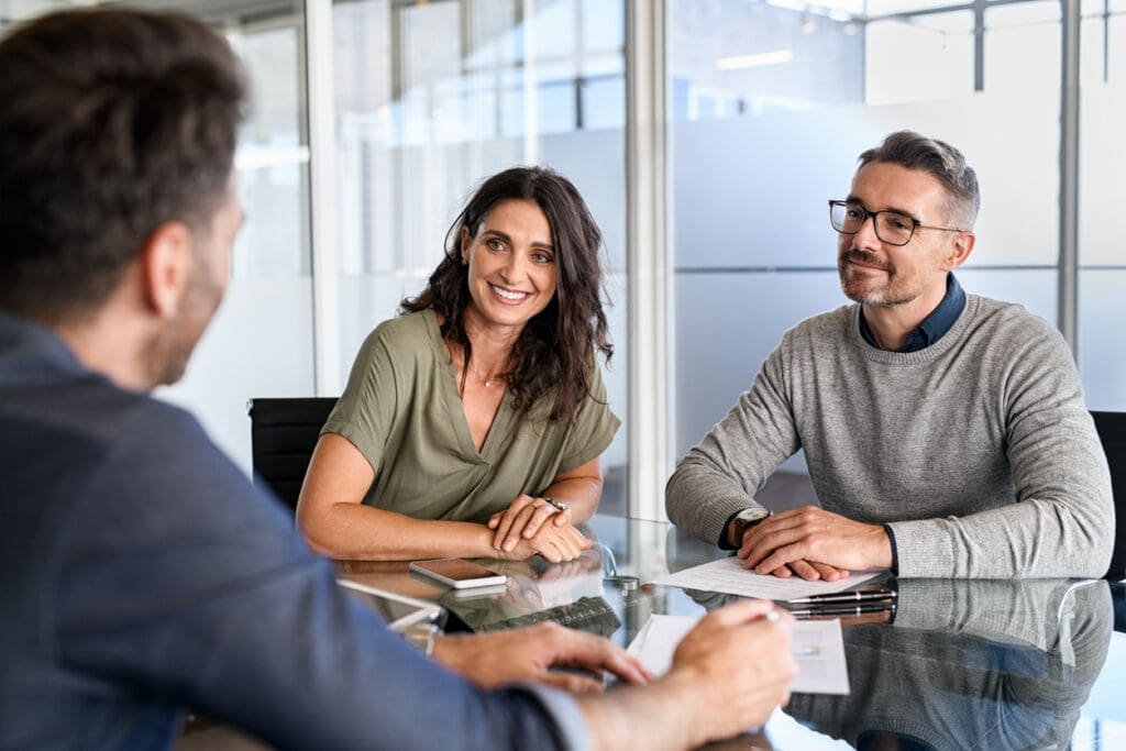 A woman and a man sit at a glass table, smiling and listening to another person in a modern office setting. Papers, a phone, eyeglasses, and cashless payments brochures are on the table.