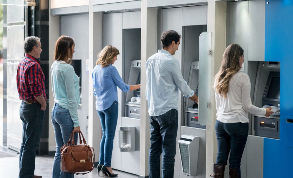 Five people stand in line at a row of Reverse ATMs; three are using the machines for digital transactions while two wait behind them. The setting appears indoors, and each person faces an ATM screen.