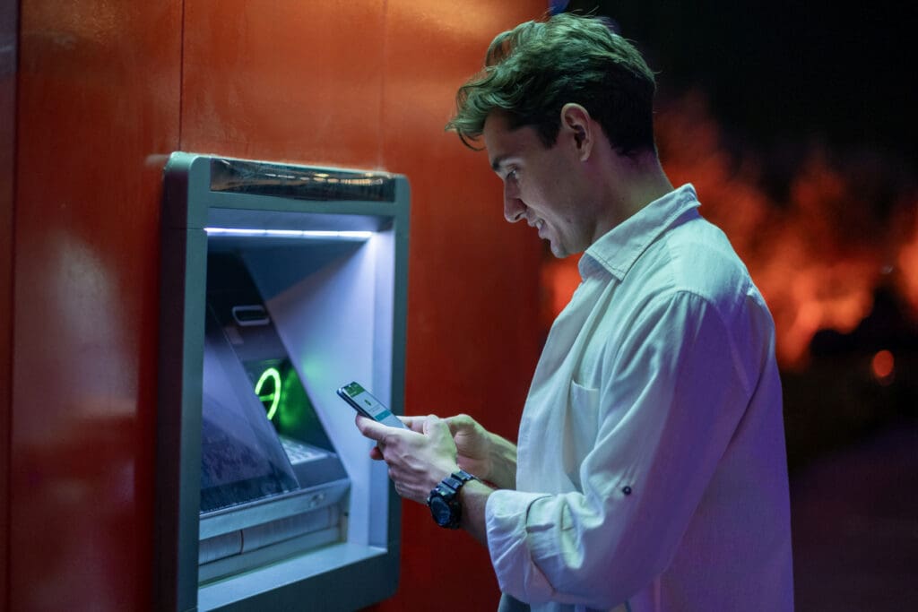 A man in a white shirt stands at a cash machine at night, looking at his smartphone, possibly checking information or confirming a transaction. The ATM glows with blue light, and the background is softly lit.