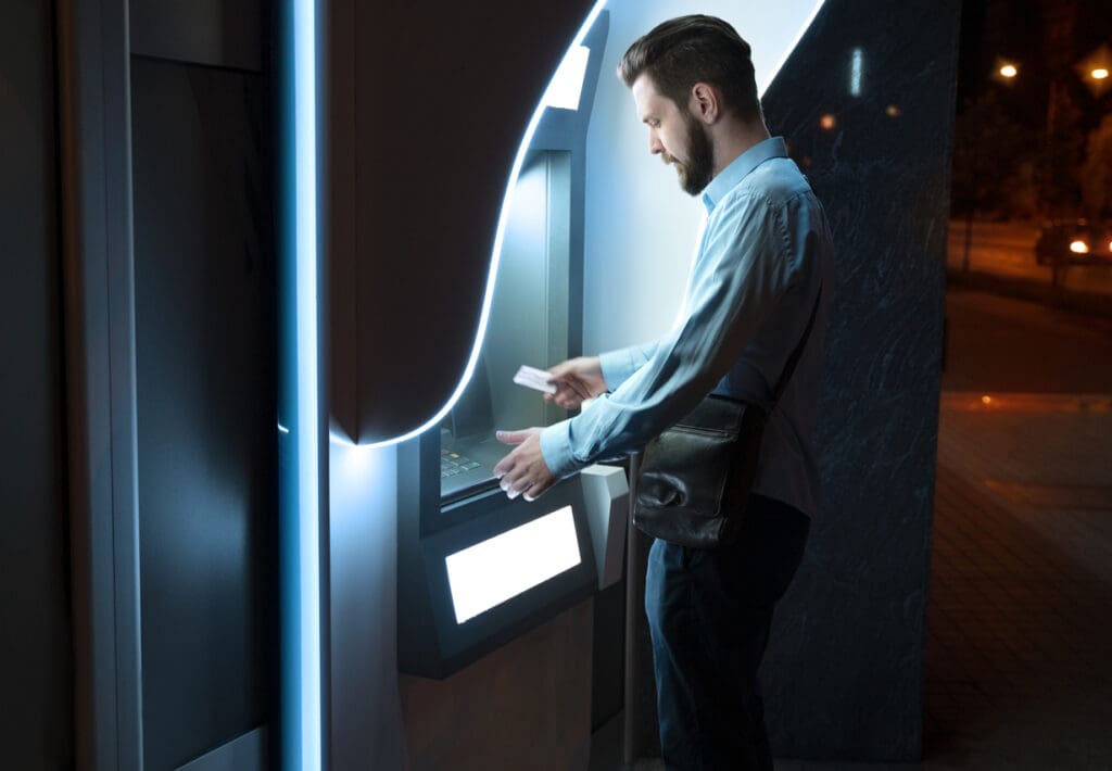 A man stands at a cash machine at night, holding a bank card and using the touchscreen. He is wearing a light blue shirt, dark pants, and a crossbody bag. The illuminated area suggests convenience for business owners seeking to maximize profits.