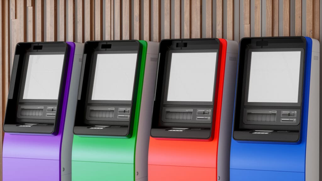 Four modern reverse ATMs in purple, green, red, and blue are lined up in front of a wooden wall, each with a blank screen and card slot visible—showcasing the latest in cashless payments and payment technology.