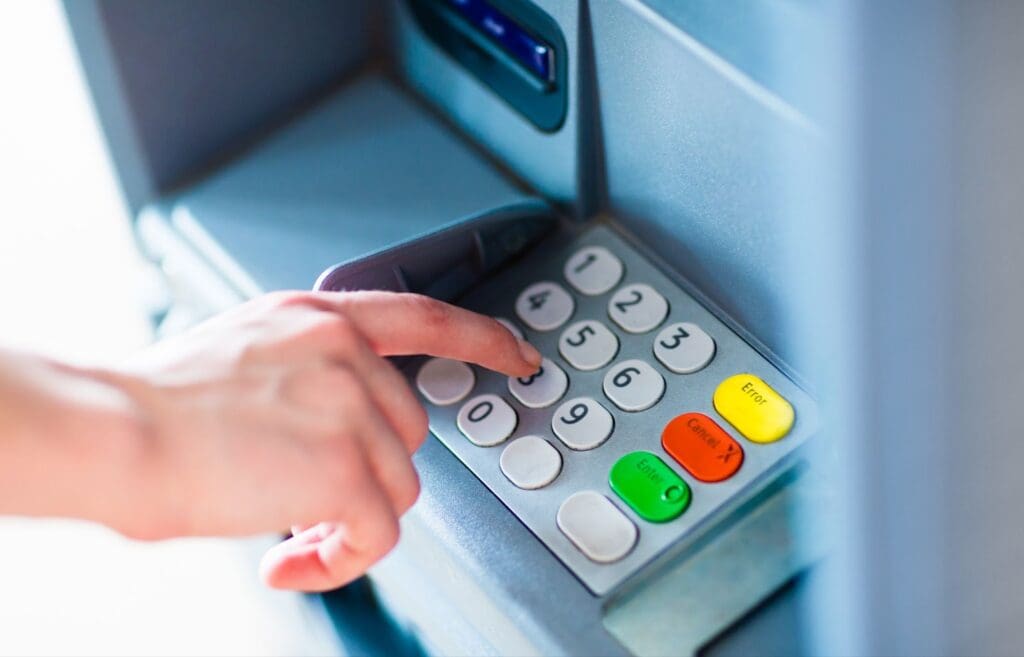 A person’s hand pressing buttons on an ATM keypad, entering a PIN or transaction amount as part of seamless payment solutions. The keypad has number and function keys, including green, yellow, and red buttons.