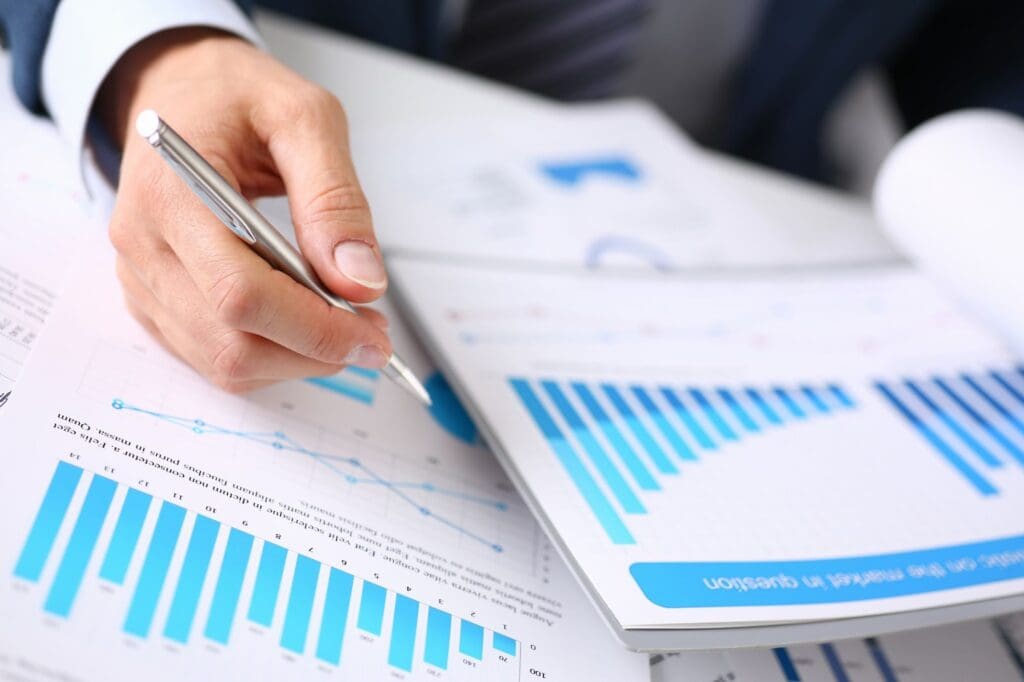 A person holding a pen reviews financial documents and charts with blue bar and line graphs, indicating data analysis or business performance review, possibly considering ATM investment opportunities.