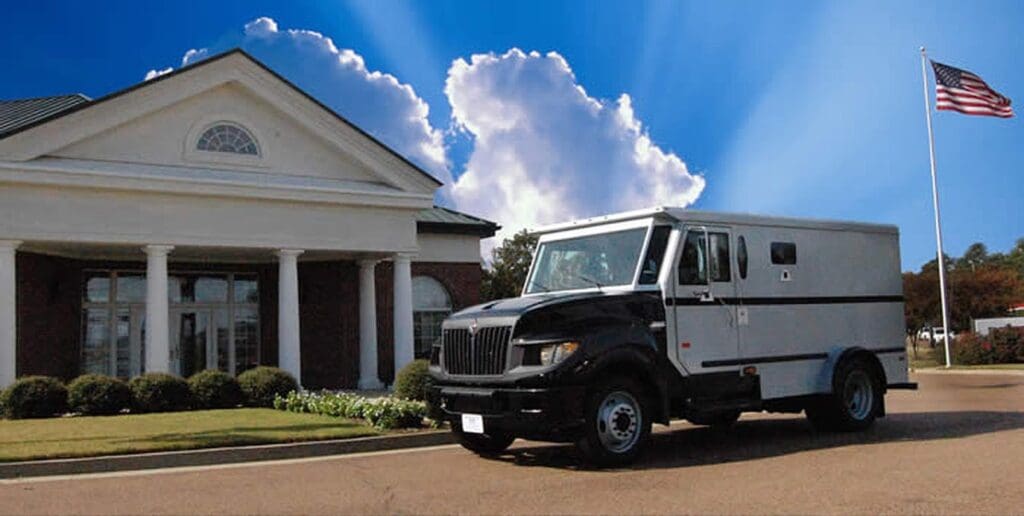 A large armored truck, used by ATM service providers, is parked on a driveway in front of a building with white columns. The sky is bright blue with clouds, and an American flag flies on a flagpole nearby.