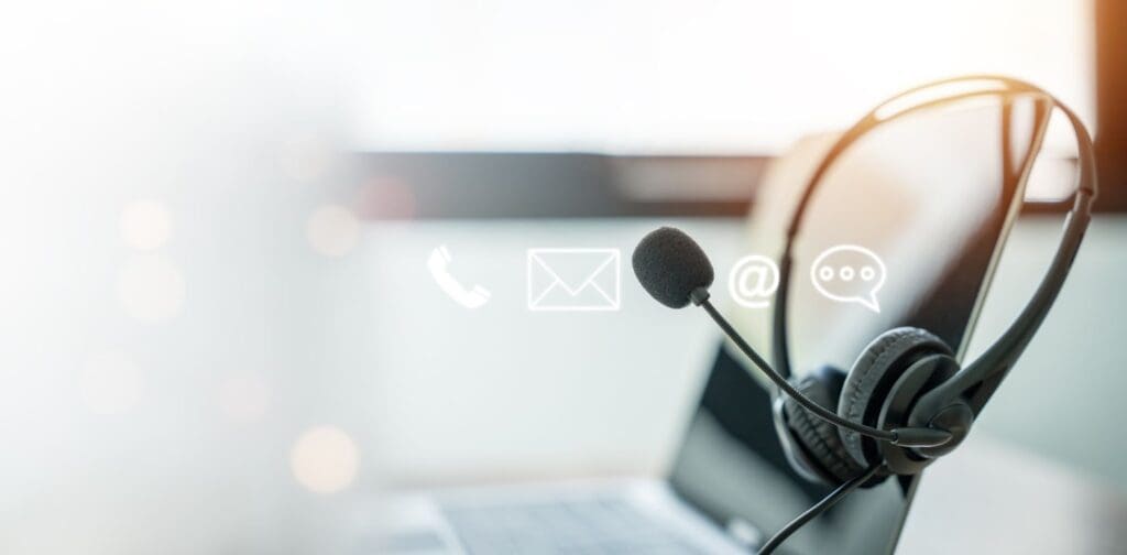 A headset rests on a laptop with icons for phone, email, and chat overlaid, suggesting customer service or ATM managed services support communication. The background is softly lit and blurred.