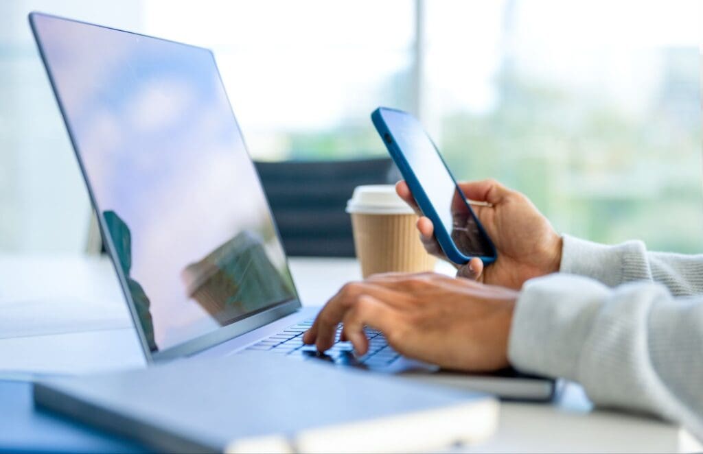 A person uses a laptop and smartphone at a desk, perhaps researching ATM service providers. A coffee cup and closed notebook are nearby. The scene is bright with natural light streaming through a window.