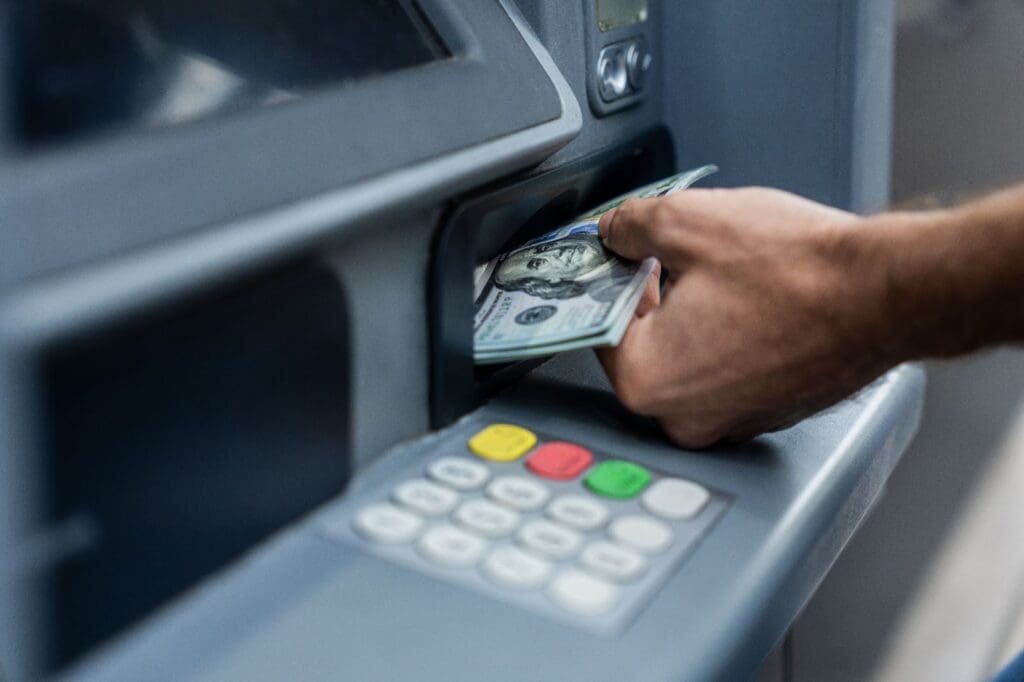 A person withdrawing several U.S. $100 bills from an ATM, their hand pulling out cash above a keypad with colorful buttons—highlighting the convenience enabled by leading ATM service providers and modern payment solutions.