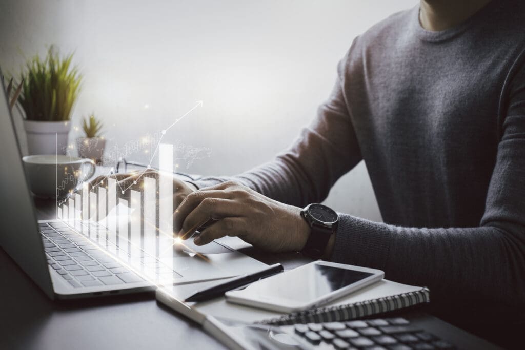 A person types on a laptop at a desk with a smartphone, notebook, calculator, and plant. A digital bar graph with an upward trend is superimposed, reflecting growth or business success for ATM service providers.