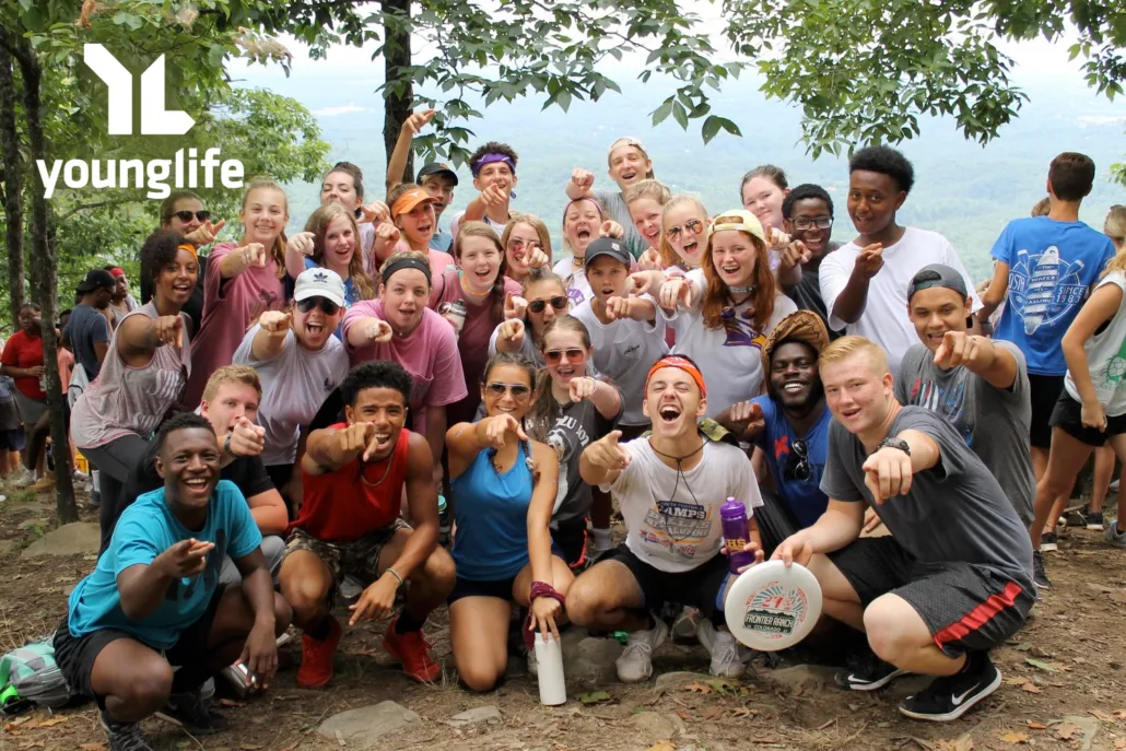 A large group of smiling teenagers and young adults pose outdoors on a hiking trail, many pointing at the camera. Trees and mountains are visible in the background. A younglife logo is in the top left corner.