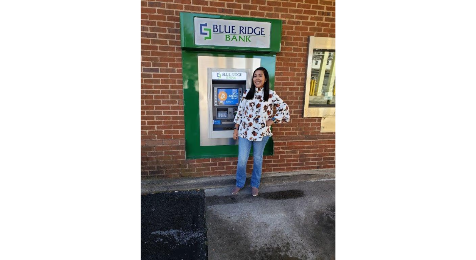 A woman in a floral shirt and jeans stands and smiles in front of a Blue Ridge Bank ATM set into a brick wall.