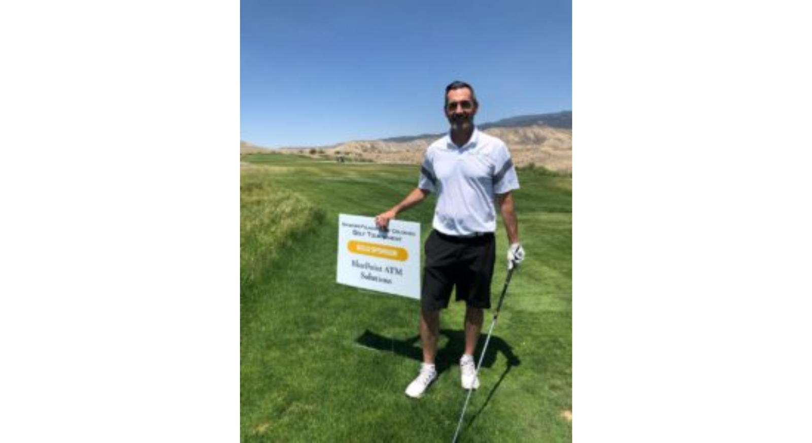 A man in a white polo shirt and black shorts stands on a golf course holding a golf club, smiling beside a sign about the event. The landscape is grassy with hills in the background under a clear sky.