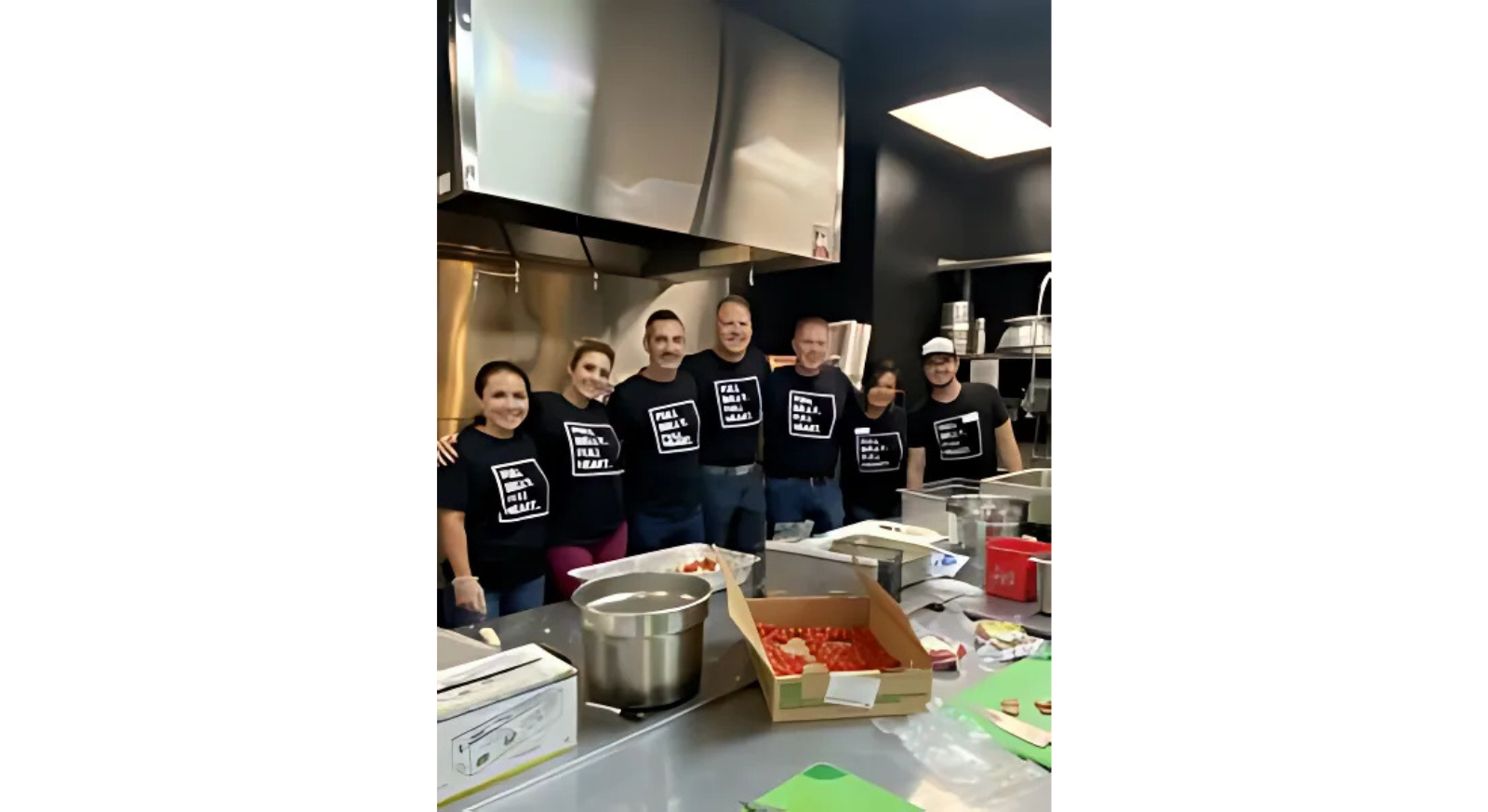 Six people wearing matching This is what REAL E.A.T. looks like! shirts stand and smile together in a commercial kitchen, surrounded by food prep stations and cooking equipment.