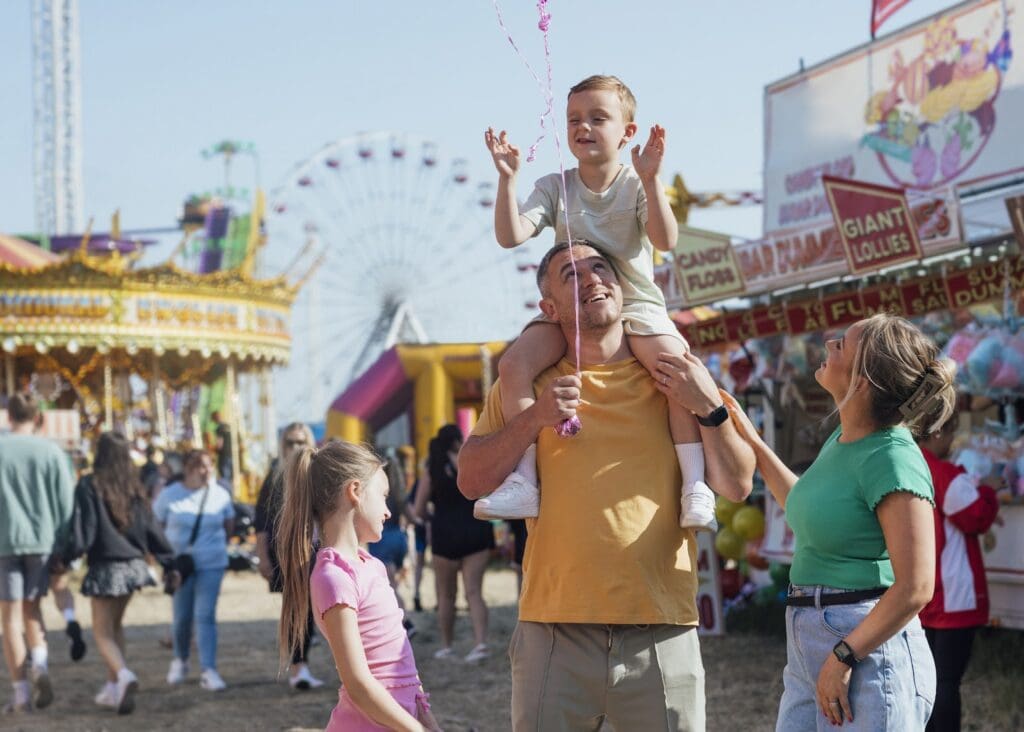 A family enjoys a day at a fairground; a man carries a boy on his shoulders while a woman and girl smile nearby. Carnival rides, booths, reverse ATMs, and a Ferris wheel can be seen in the background.