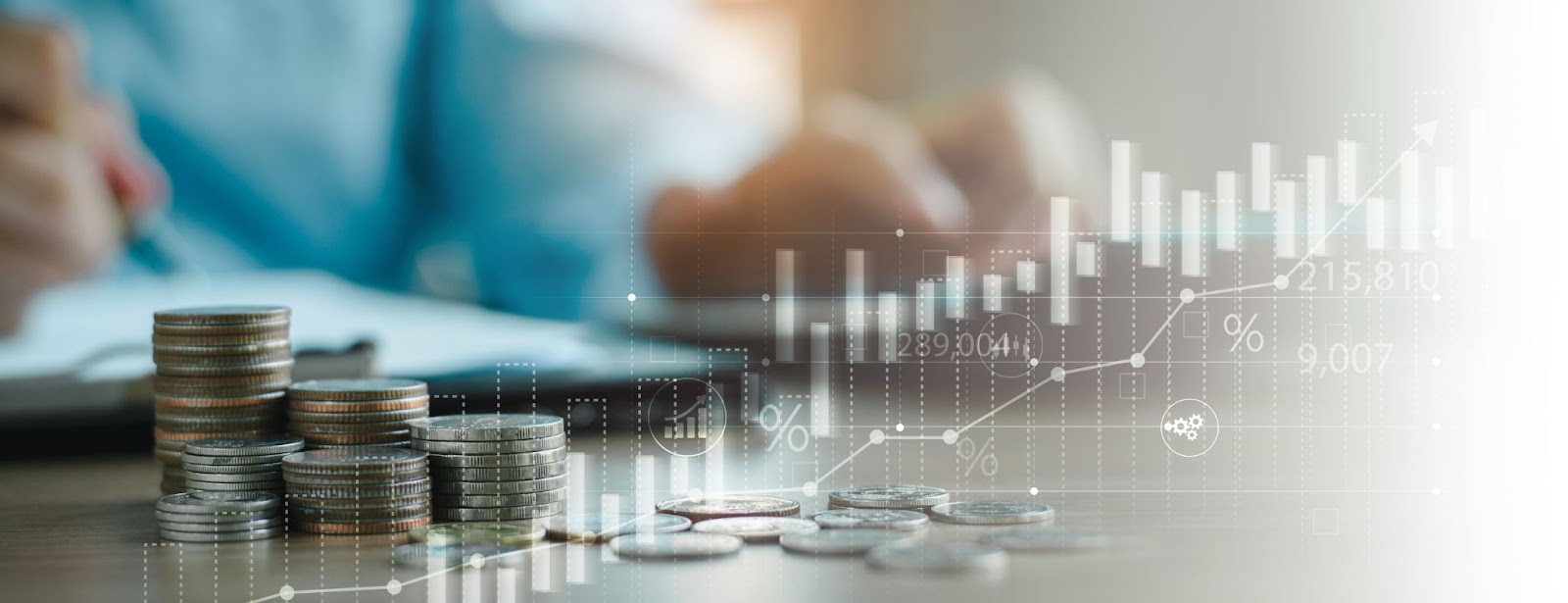 Stacks of coins and scattered coins on a table, with a blurred person writing in the background. Overlay of financial graphs and data, suggesting growth, investment, or analysis by the best ATM service providers.