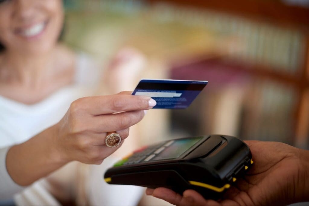 A smiling woman holds a prepaid card near a payment terminal, making a contactless payment. The background is blurred, focusing on her hand and the card reader—ideal for settings using Reverse ATMs or prepaid card machines.