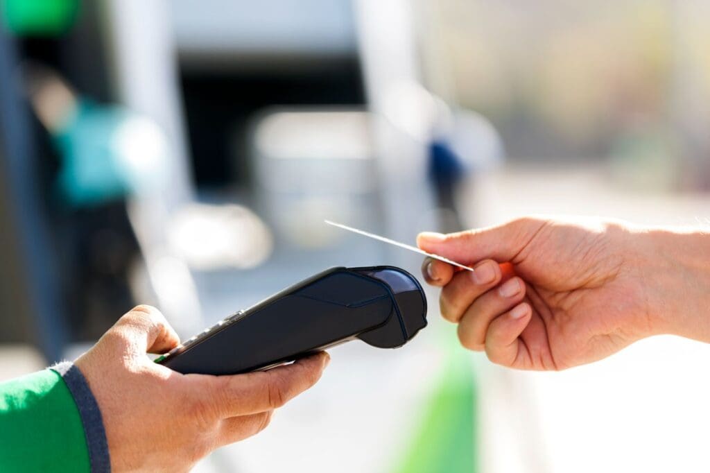 A person holds a payment terminal, similar to those used at reverse ATMs, while another holds a card near it for a contactless payment outdoors. The background is blurred.