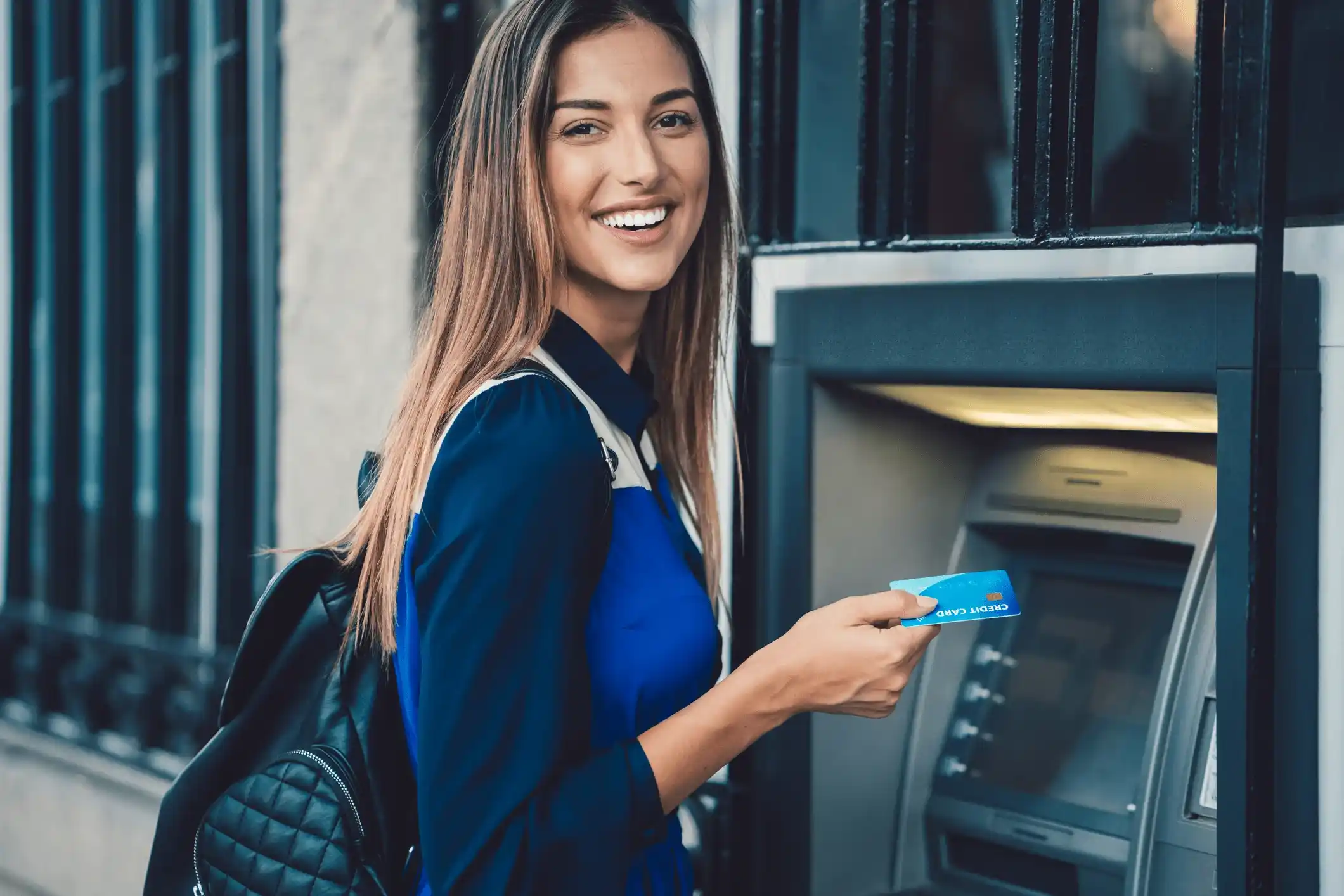 A smiling woman with long brown hair, wearing a blue shirt and carrying a black backpack, stands at an ATM holding a bank card.
