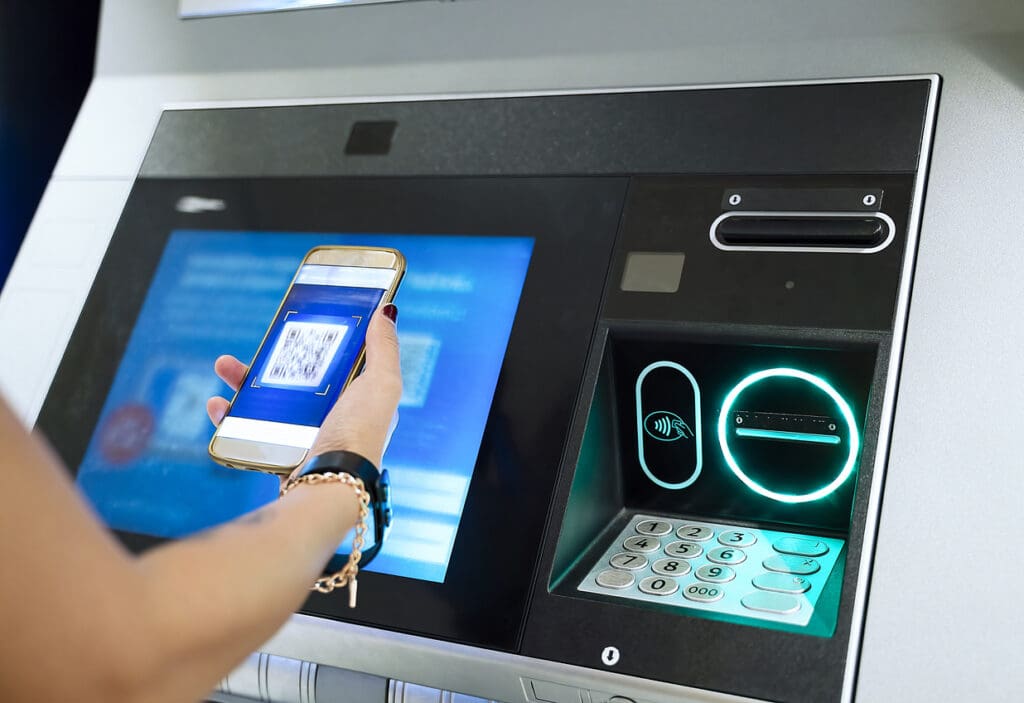 A person holds a smartphone displaying a QR code near an ATM screen, preparing to use contactless or mobile banking features at the machine—an increasingly common scene as reverse ATMs become more popular.