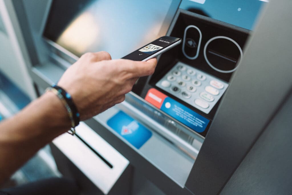 A person holds a smartphone near an ATM keypad, possibly using contactless payment or authentication, with their arm visible and the ATM screen and card slot in the background, highlighting the growing use of reverse ATMs for cashless transactions.