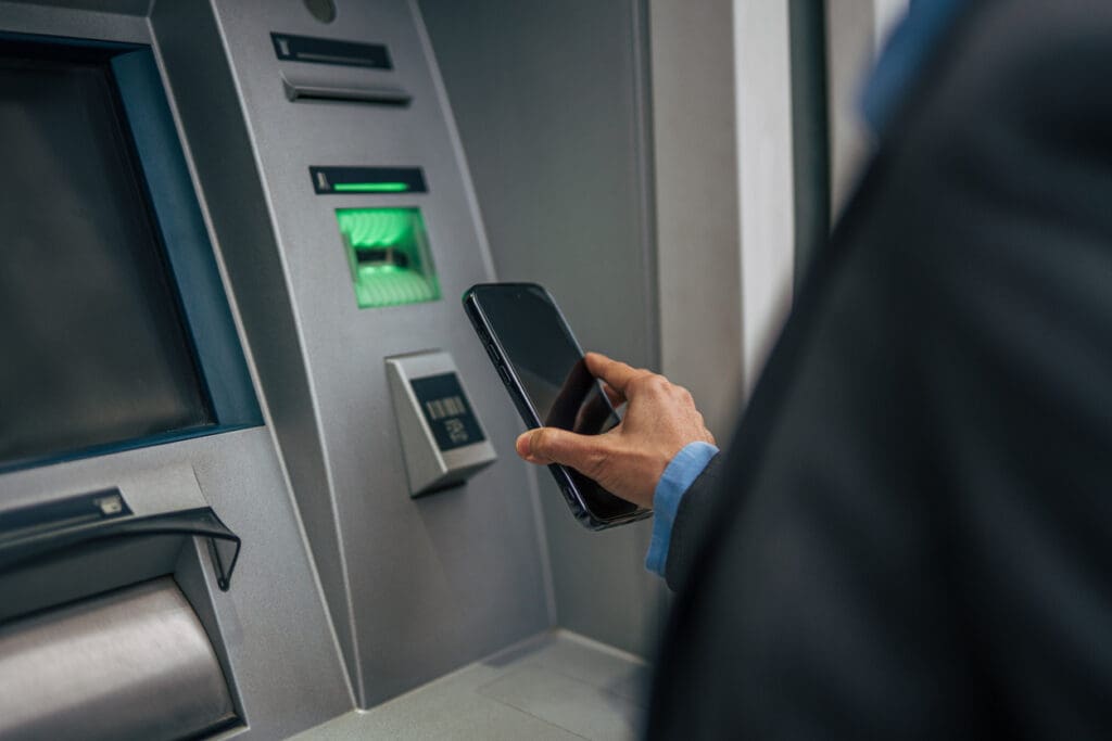 A person holds a smartphone near an ATM, possibly using contactless technology for a transaction. The ATM screen, card reader, and reverse ATMs feature are visible, with the person’s hand in focus.
