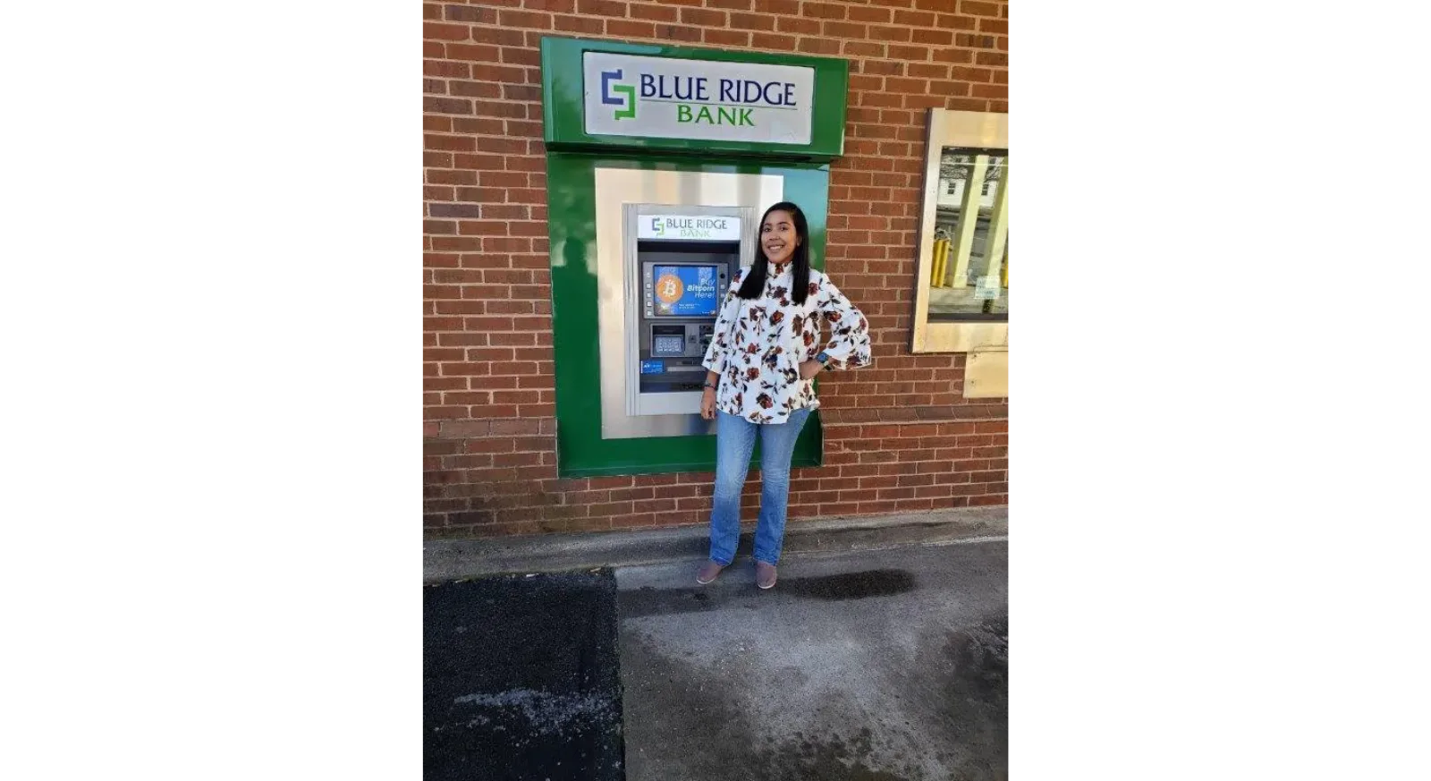 A woman stands and smiles in front of a Blue Ridge Bank ATM attached to a brick wall. She is wearing a white floral top, blue jeans, and pink shoes.