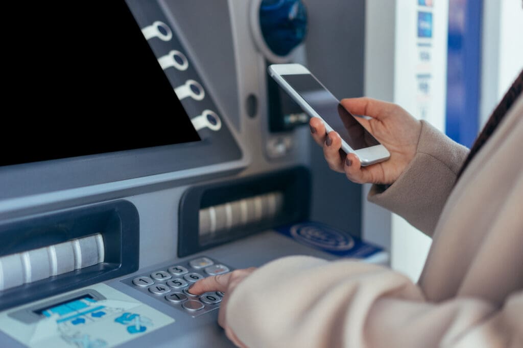 A person wearing a beige coat holds a smartphone in one hand while using the keypad on an ATM with the other, highlighting how ATMs remain relevant alongside digital payments. The ATM screen is blank.