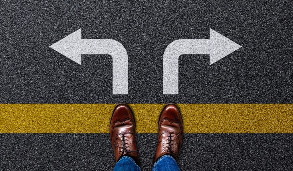 A person wearing brown shoes and blue jeans stands at a yellow line on asphalt, facing two large white arrows pointing left and right—much like choosing between ATM vendors, each presenting a different direction or opportunity.