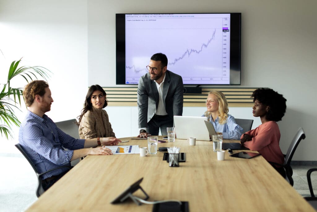 Five people in business attire meet around a conference table, with a man standing and smiling. A large screen behind them displays a line graph trending upward as they discuss reverse ATMs. Laptops and papers are on the table.