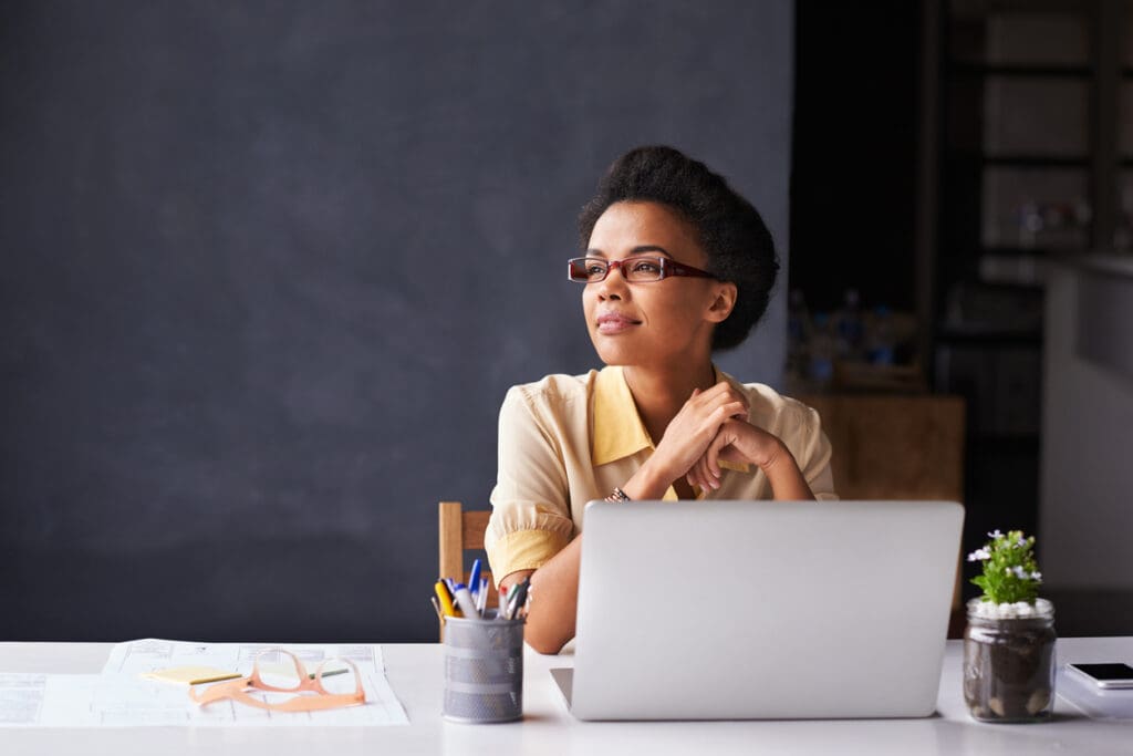 A woman wearing glasses sits at a desk with a laptop, papers, and stationery, looking thoughtfully to the side. Alongside a small potted plant and a chalkboard, a pamphlet about reverse ATMs is visible on the desk.