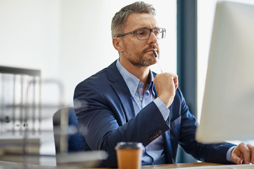 A man in a blue suit and glasses sits at a desk, thoughtfully looking at a computer screen while holding a pen to his mouth. A takeaway coffee cup and information about reverse ATMs are on the desk in front of him.