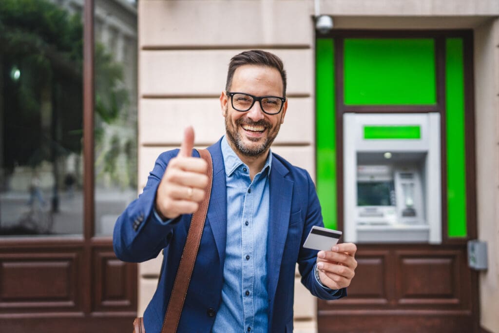 A smiling man in a blue suit and glasses stands in front of a reverse ATM, holding a credit card in one hand and giving a thumbs-up with the other.