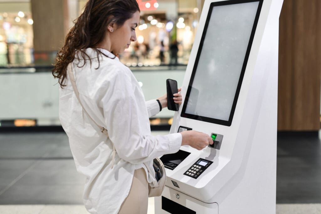 A woman uses a touchscreen cash-to-card kiosk in a public indoor space, holding her phone and pressing a button to enjoy convenient payment benefits.