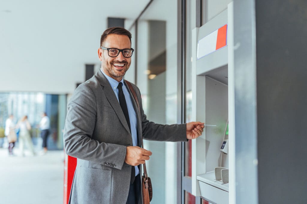 A smiling man in a suit and glasses uses a reverse ATM outdoors, holding a card in his hand. He stands beside the machine, looking at the camera, with a modern building in the background.