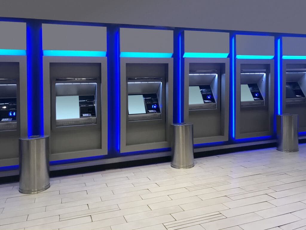 A row of modern reverse ATMs with blue lighting, metal trash bins in front, and a white tiled floor in a clean, indoor setting.