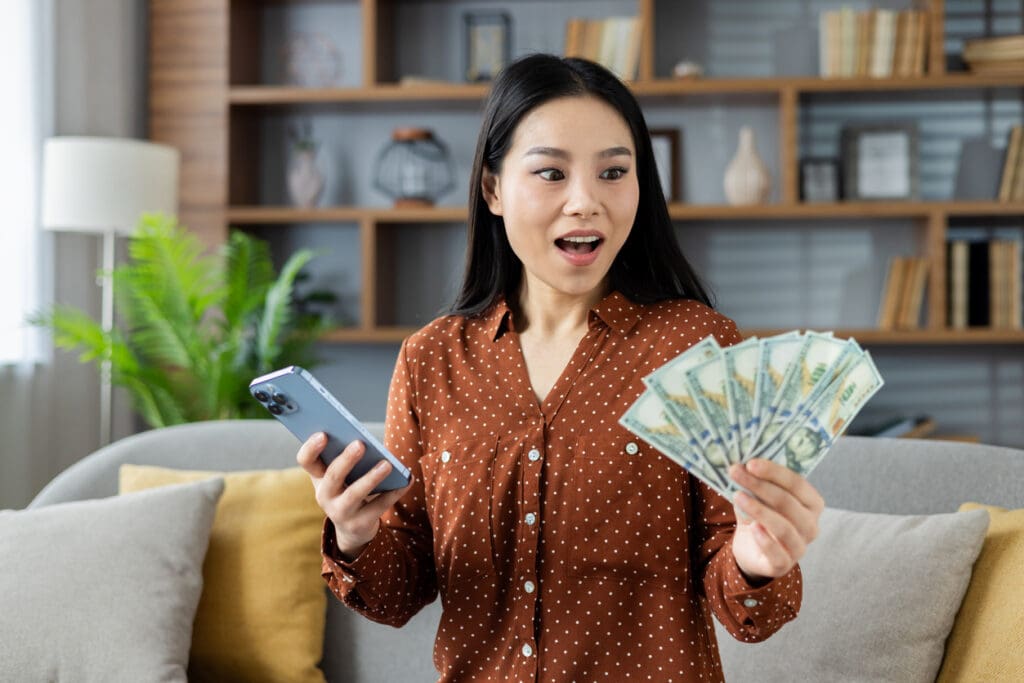 A woman holding a smartphone in one hand and fanned-out U.S. dollar bills in the other looks surprised and happy while standing in a modern living room with bookshelves in the background.