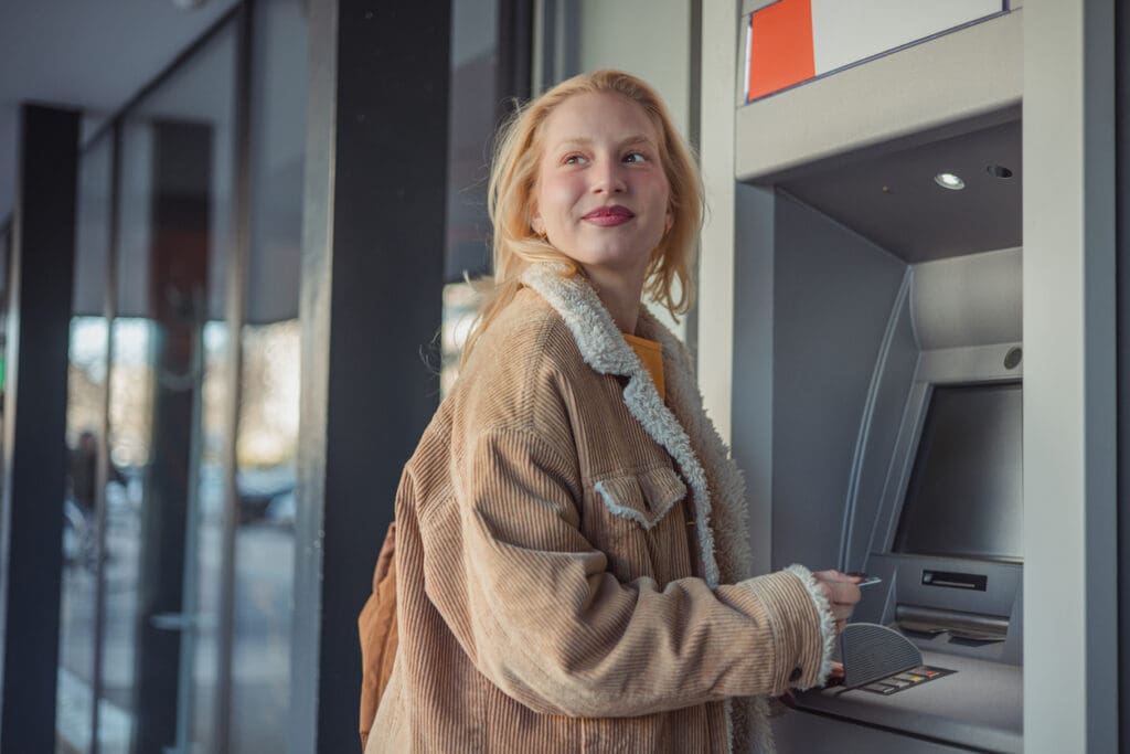 A young woman in a tan corduroy jacket uses an outdoor Prepaid Card ATM, looking to the side and smiling while holding a bank card in her hand.