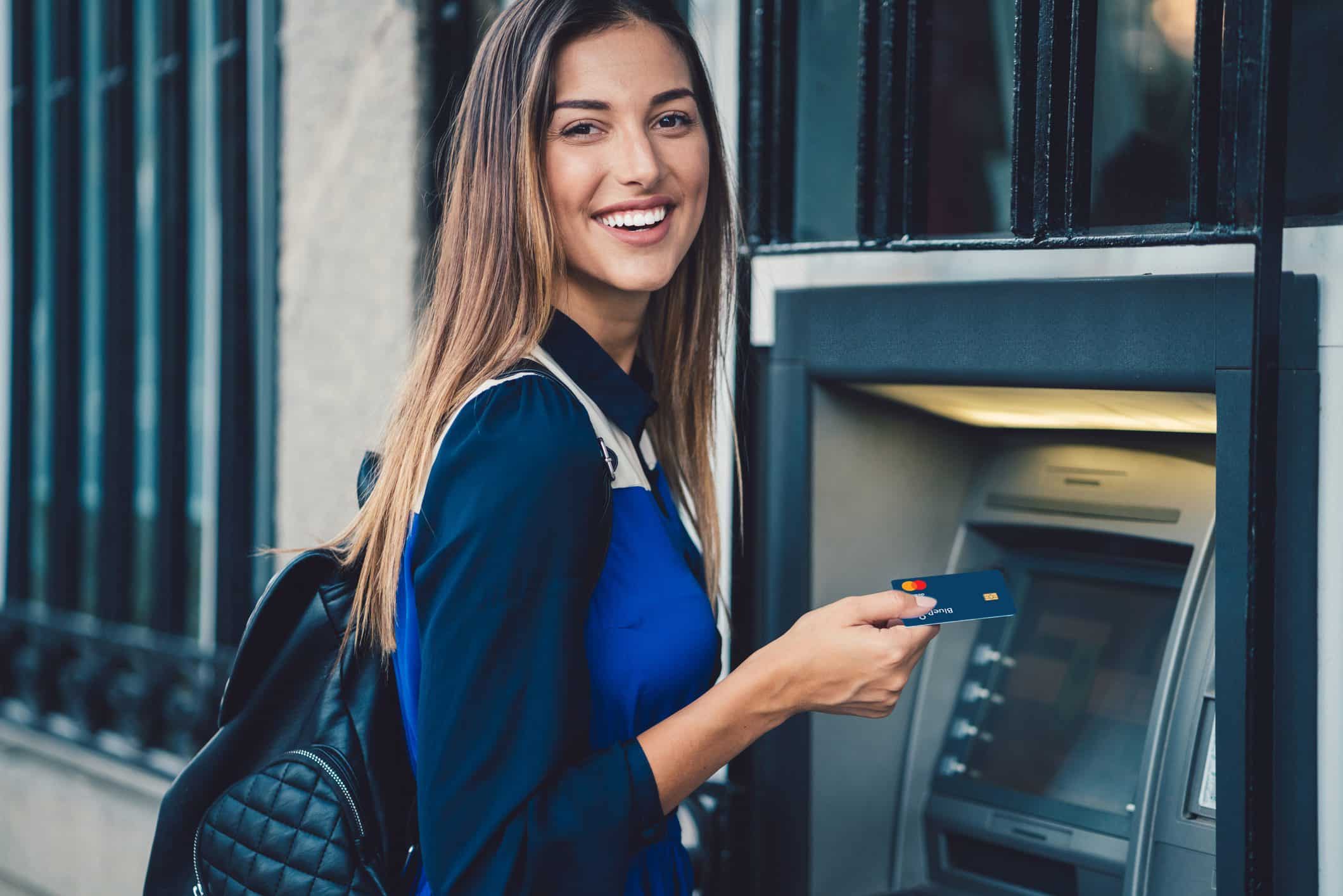 A smiling woman in a blue shirt stands at an ATM in New York, holding a credit card in her hand and wearing a black backpack, reflecting the city’s shift toward cashless transactions ahead of the compliance deadline.